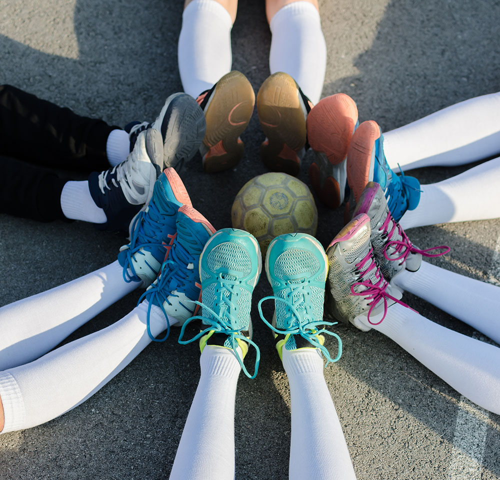 Vertical image of young female players of handball sitting on court with stretched legs and ball in the middle.